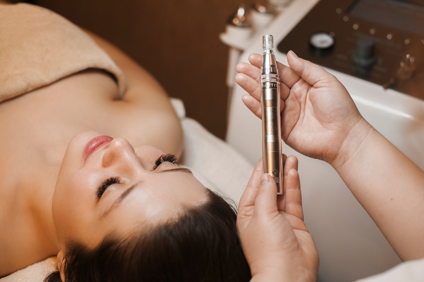 Upper view of a cosmetologist hands holding a dermapen for future microneedling procedure on womans face which is leaning on a bed with closed eyes.